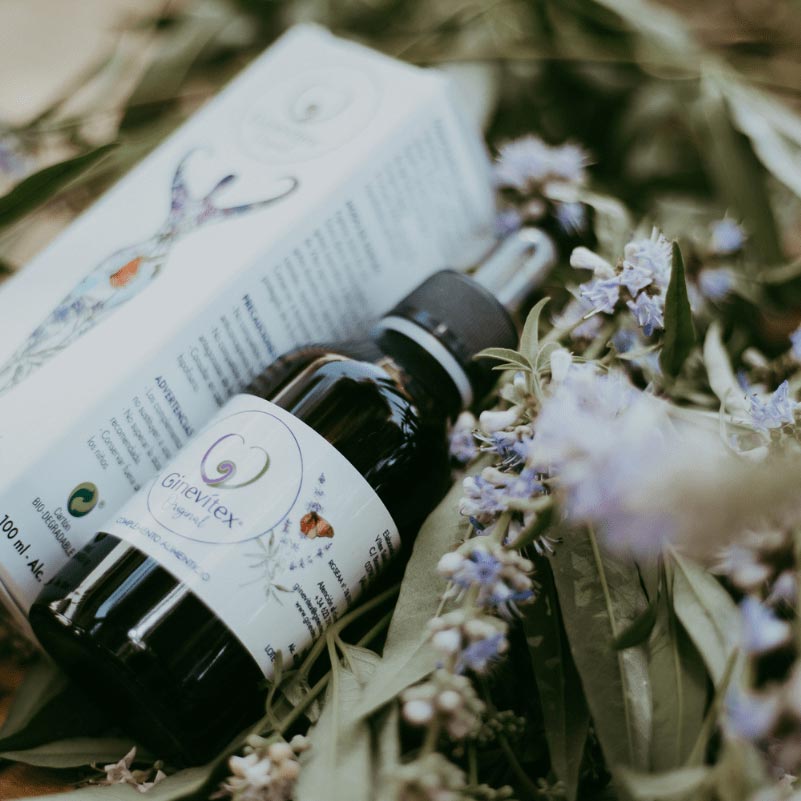 A 100ml bottle of Ginevitex food supplement laying on dry flowers from the plant Vitex agnus castus laying in front and the package box on the side