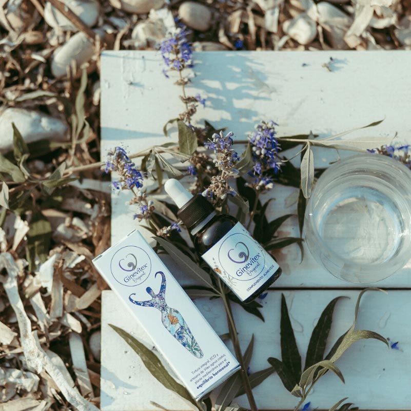A 30ml bottle of Ginevitex food supplement laying on a table with branches and flowers of the plant Vitex agnus castus . The package box is laying beside and a glass of water on the right side.