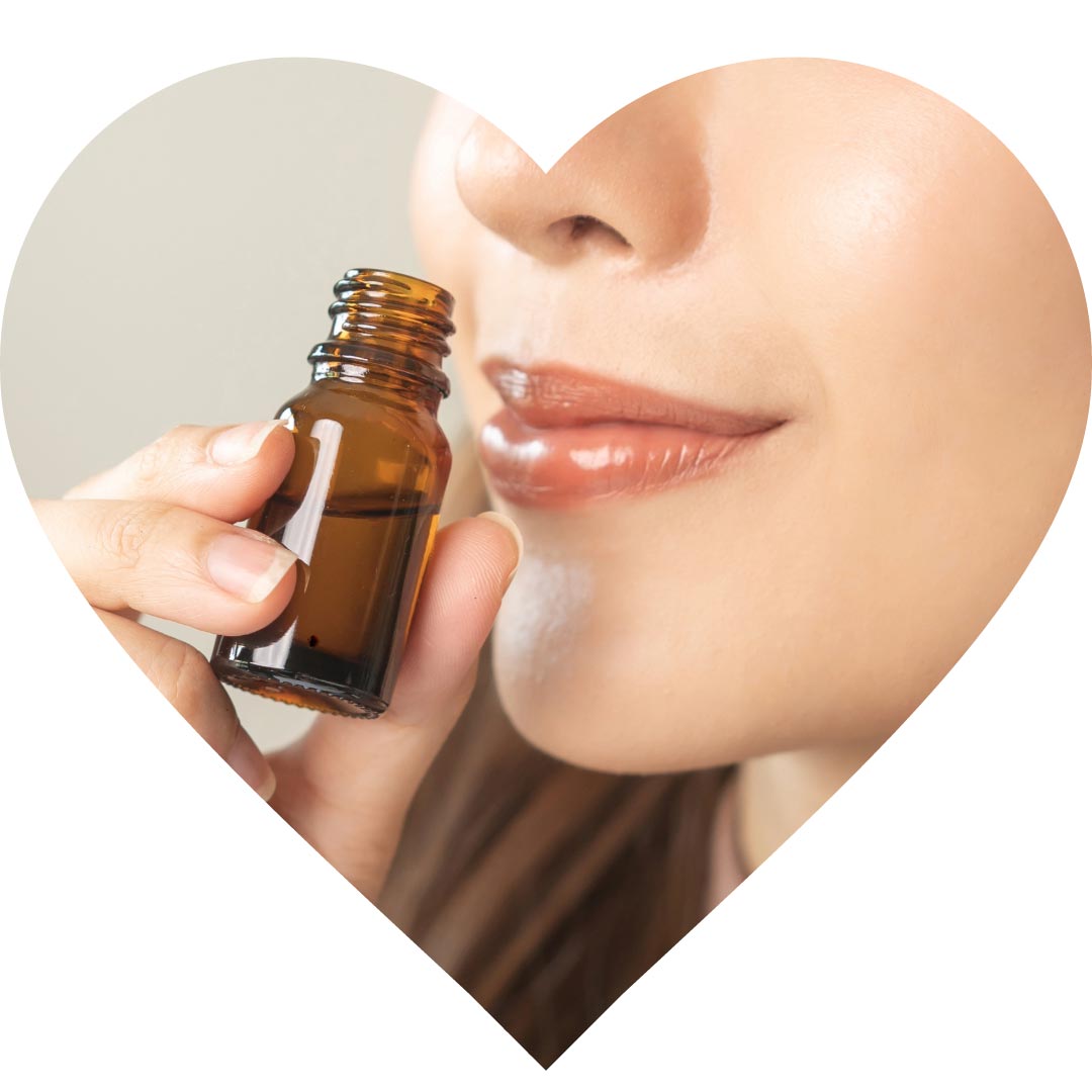 A woman inhaling essential oil from a brown glass bottle, the image is in the shape of a heart