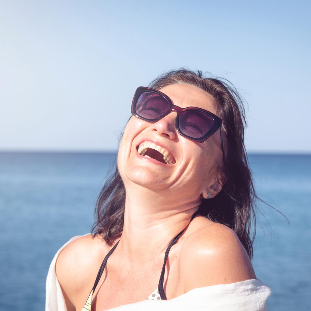 A brunette woman laughing with her head back, wearing a bikini top with a shirt over the shoulders and sunglasses. In the background is the sea