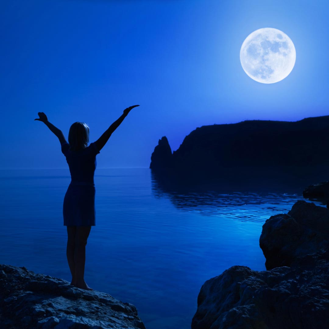 Woman standing in the dark by the see, greeting the full moon