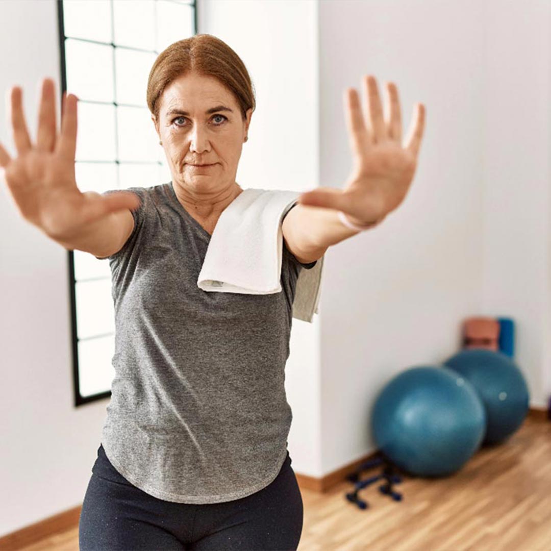 A woman in her middle age raising her hand as in stop with a towel over her shoulder, wearing work out clothes and in the background pilates balls