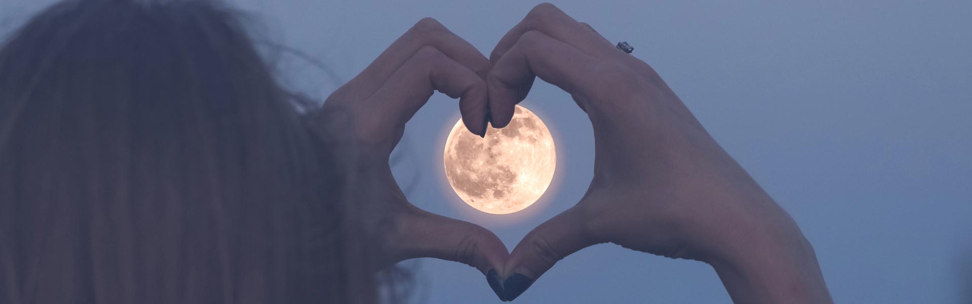A woman holding her hands in shape of a heart around the full moon on the sky at night