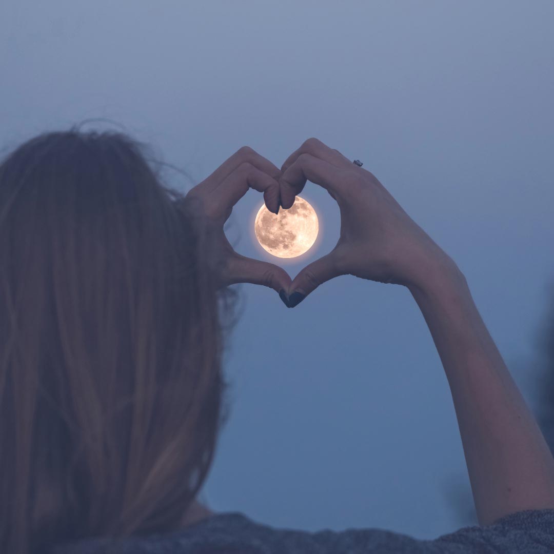 A woman holding her hands in shape of a heart around the full moon on the sky at night